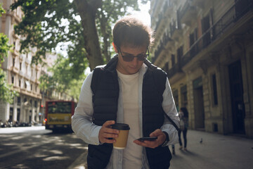 Coffee on a go. Caucasian young businessman in casual clothes and sunglasses checking news via mobile phone with takeaway paper coffee cup. Communication outdoors with gadget