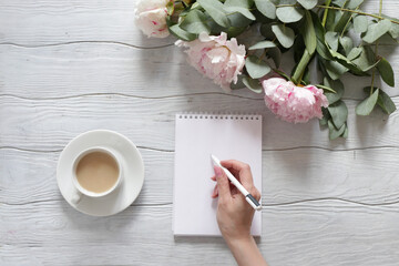 A woman's hand writes in a notebook. Table with peonies and a Cup of coffee top view.
