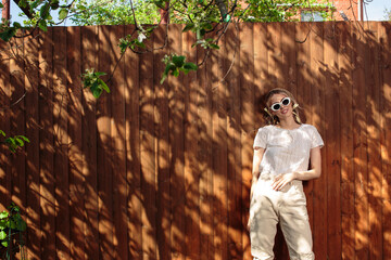 Portrait of a pretty girl in glasses and a headscarf standing against a wooden fence in the backyard on a Sunny day.