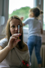 Mother is showing syringe while daughter is looking through window.Shot of young mother holding her medication.Cute girl turned back to her mother,looking out the window,while mother takes medication