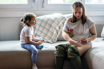 Young mother sitting on couch and giving her self injection, while her daughter watching her. Women...