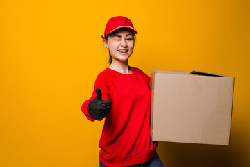Young asian delivery woman holding box and thumbs up on yellow background