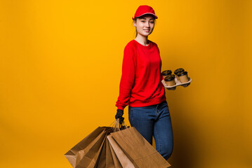 Young delivery woman holding coffee and papers bags isolated on yellow background