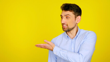 Handsome bearded man in light blue shirt smiles and shows with hand place for products and text. The guy on yellow background