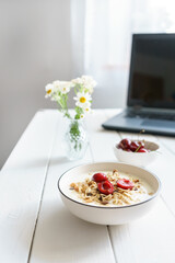 müsli mit frischen Kirschen auf einem weißen Holztisch mit einem Laptop im Hintergrund und frischen Blumen in einer Vase. Home office, gesundes Frühstück.