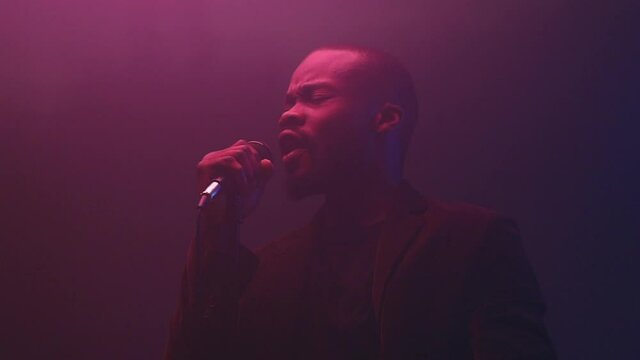 a dark-skinned handsome guy with a beard  in dark jacket and t-shirt holds a microphone in his hand and emotionally sings in a dark studio with neon light