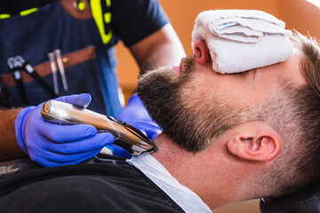 Hipster sitting in the barber's room. The barber touches up his beard with an electric cutter. ...