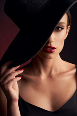portrait of a beautiful tanned girl with professional makeup, red lips, on a burgundy background in a black dress with straps and black hat that covers her eye, she looks at camera with serious face