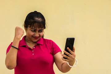 Indian female model looking at mobile phone happily with slight smiling face in yellow background with copy space for text