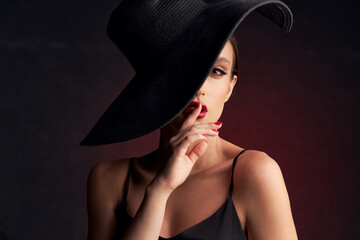 portrait of beautiful tanned girl with professional makeup, red lips, on a burgundy background in a black dress with straps and black hat that covers her eye, she looks at the camera touching her lips
