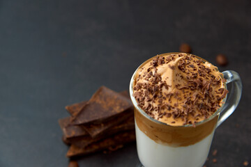 Glass of trendy fluffy creamy dalgona or whipped instant coffee with milk, coffee beans and chocolate on the dark background. New popular food and drink trend concept. Copy space.