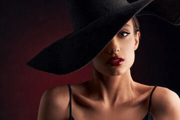 portrait of beautiful tanned girl with professional makeup, red lips, on a burgundy background in a black dress with straps and black hat that covers her eye, she looks at the camera with serious face