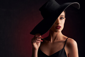 portrait of a beautiful tanned girl with professional makeup, red lips, on a burgundy background in a black dress with straps and black hat that covers half of her face and she looks at the camera