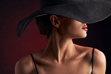 profile portrait of a beautiful tanned girl with professional makeup, red lips on a burgundy background in a black dress with straps, a black hat that covers half of her face