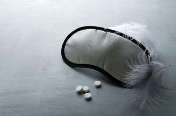 Concept of relaxation and deep sleep.Sleep mask, white tablets and feathers on the grey...