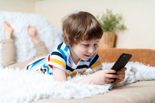 Happy little child boy playing online game, watching video on cellphone, lying on sofa entertaining in living room. Smiling small kid using funny mobile apps, enjoying free leisure time at home.