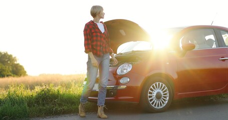 Portrait of cheerful blonde hipster girl having problem with car on roadtrip. Woman near the broken car with open hood
