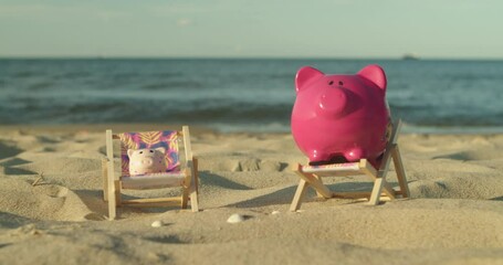Two piggy banks sit on mini sun loungers on the sandy beach. A man's hand throws a coin into a piggy bank. Male Hand Throws a Coin Into a Piggy Bank a Pig on a Sandy Shore. Slow Motion. - Powered by Adobe