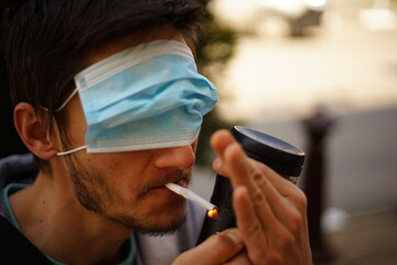 A portrait of a man with a face mask on his eyes. He is lighting a cigarette.
