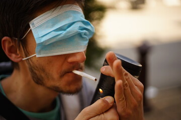 A portrait of a man with a face mask on his eyes. He is lighting a cigarette.
