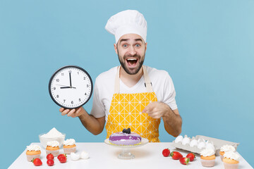 Excited male chef or cook baker man in apron white t-shirt toque chefs hat cooking at table isolated on blue background. Cooking food concept. Mock up copy space. Hold clock pointing finger on camera.