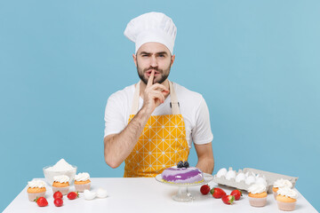 Secret young male chef or cook baker man in apron white t-shirt toque chefs hat cooking at table isolated on blue background. Cooking food concept. Say hush be quiet with finger on lips shhh gesture.