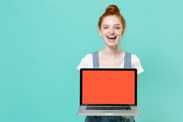 Naklejka premium Cheerful young readhead girl in casual denim clothes posing isolated on blue turquoise background studio. People lifestyle concept. Mock up copy space. Hold laptop pc computer with blank empty screen.