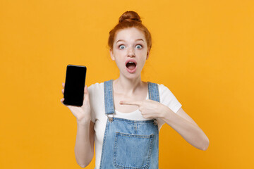 Shocked young readhead girl in casual denim clothes white t-shirt isolated on yellow background studio portrait. People lifestyle concept. Point index finger on mobile phone with blank empty screen.