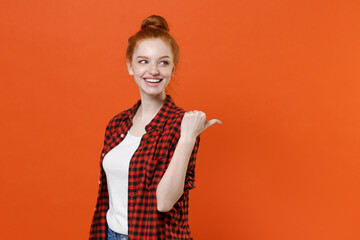 Smiling young readhead girl in casual red checkered shirt posing isolated on orange background studio portrait. People sincere emotions lifestyle concept. Mock up copy space. Pointing thumb aside.