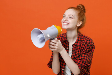 Smiling young readhead girl in casual red checkered shirt posing isolated on orange background studio portrait. People lifestyle concept. Mock up copy space. Screaming on megaphone, looking aside.