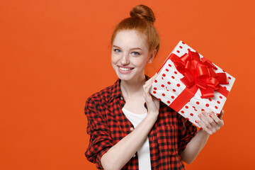 Smiling young readhead girl in checkered shirt posing isolated on orange background. St. Valentine's Day International Women's Day birthday holiday concept. Hold red present box with gift ribbon bow.