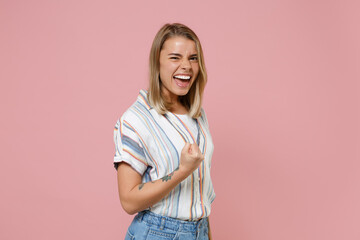 Cheerful young blonde woman girl in casual striped shirt posing isolated on pastel pink background studio portrait. People sincere emotions lifestyle concept. Mock up copy space. Doing winner gesture.