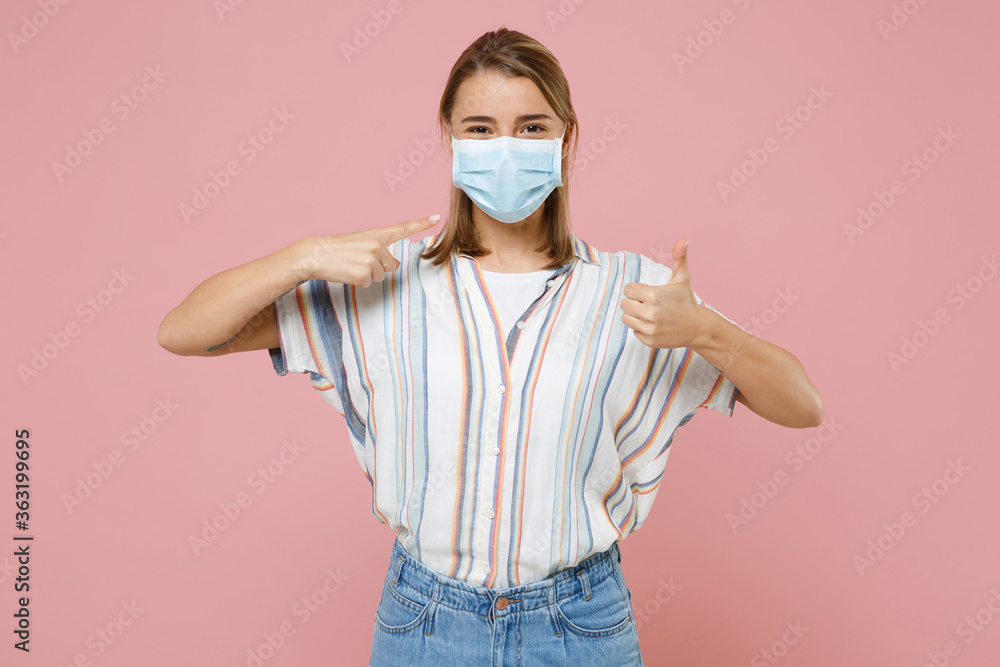 Wall mural Young blonde girl in striped shirt isolated on pink background. Epidemic pandemic coronavirus 2019-ncov sars covid-19 flu virus concept. Pointing index finger on sterile face mask, showing thumb up.