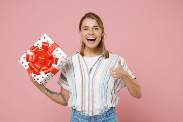 Funny young blonde woman girl in striped shirt isolated on pastel pink background. Valentine's Day Women's Day birthday, holiday concept. Hold red present box with gift ribbon bow, showing thumb up.