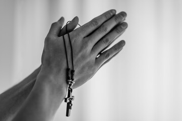 Christian male person hands praying while holding a pendant with a crucifix. Black and white cropped closeup of hands together raised up in prayer.