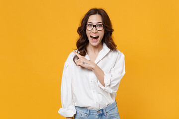 Surprised young brunette business woman in white shirt glasses isolated on yellow background studio portrait. Achievement career wealth business concept. Mock up copy space. Point index finger aside.