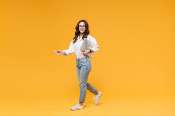 Cheerful young brunette business woman in white shirt glasses isolated on yellow background in studio. Achievement career wealth business concept. Mock up copy space. Hold laptop pc computer, walking.