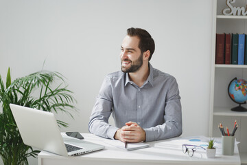 Obraz premium Smiling handsome young bearded business man in gray shirt sitting at desk, work on laptop pc computer in light office on white wall background. Achievement business career concept. Looking aside.