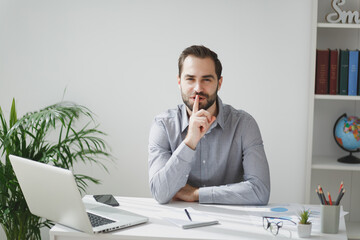 Secret young business man in gray shirt sit at desk work on laptop in light office on white wall...