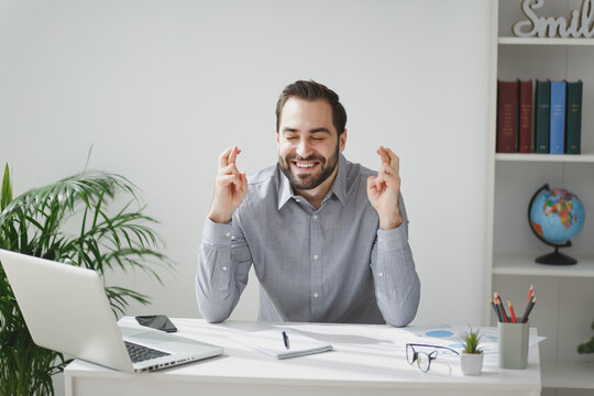 Smiling business man in gray shirt sit at desk in light office on white wall background. Achievement business career concept. Wait for special moment, keeping fingers crossed, eyes closed making wish.