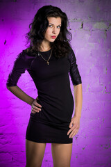 Beautiful girl in a black dress on a brick wall background. The wall is illuminated by a purple neon. A beautiful stylish brunette stands near the wall.