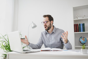 Naklejka premium Angry young bearded business man in gray shirt glasses sit at desk in light office on white wall background. Achievement business career concept. Work on laptop pc computer hold pen spreading hands.