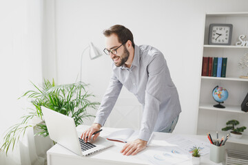 Smiling attractive young bearded business man in gray shirt glasses standing near desk in light office on white wall background. Achievement business career concept. Working on laptop pc computer.