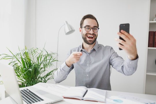 Funny business man in gray shirt sit at desk work on laptop in light office on white wall background. Achievement business career concept. Talk point index finger on on mobile phone making video call.