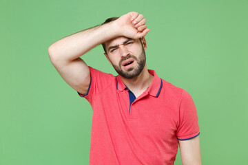 Fototapeta premium Exhausted tired young bearded man guy in casual red pink t-shirt posing isolated on green background studio portrait. People lifestyle concept. Mock up copy space. Put hands on head, having headache.