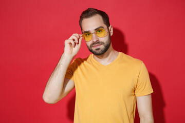 Handsome young bearded man guy in casual yellow t-shirt glasses posing isolated on red wall background studio portrait. People sincere emotions lifestyle concept. Mock up copy space. Looking camera.