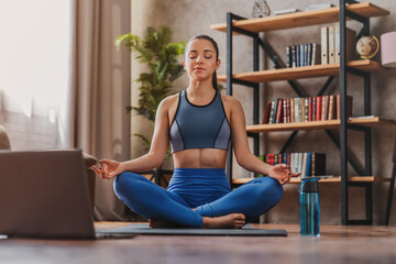 Fototapeta na wymiar Online yoga maditation home. Young girl sitting in lotus pose near laptop at home