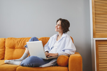 Charming young woman girl in casual clothes sitting on couch spending time in living room at home. Rest relax good mood leisure lifestyle concept. Mock up copy space. Working on laptop pc computer.