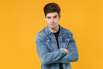 Handsome young man guy 20s in casual denim jacket posing isolated on yellow wall background studio portrait. People sincere emotions lifestyle concept. Mock up copy space. Holding hands crossed.