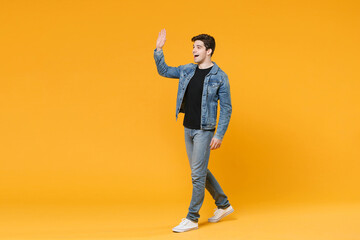 Fototapeta na wymiar Excited young man guy wearing casual denim clothes posing isolated on yellow wall background studio. People lifestyle concept. Mock up copy space. Waving and greeting with hand as notices someone.